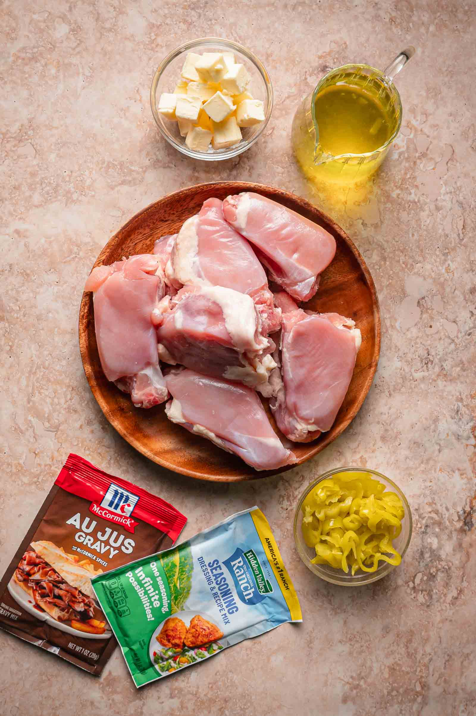 Raw chicken thighs on a plate with butter cubes, chicken broth, sliced pepperoncini, and packets of au jus gravy mix and ranch seasoning arranged on a countertop.