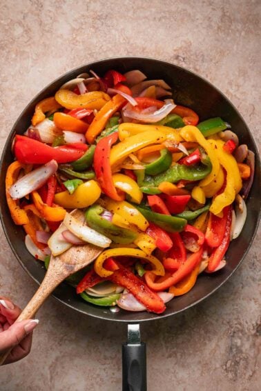 A pan of sautéed bell peppers and onions being stirred with a wooden spoon.