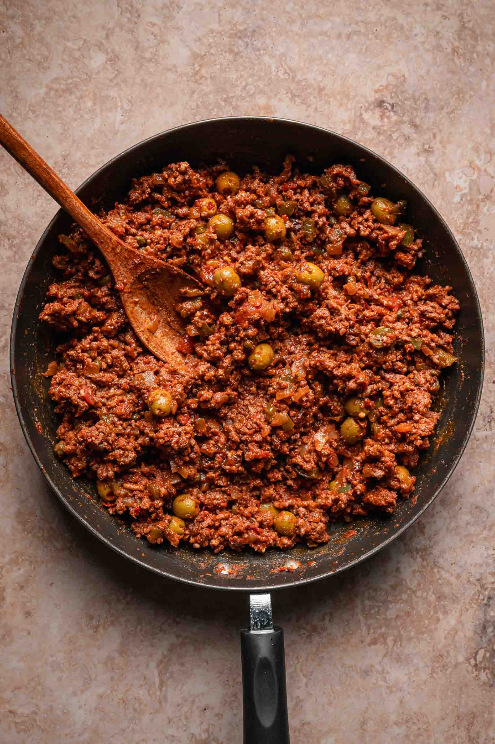 A skillet filled with cooked ground beef mixed with green olives, with a wooden spoon resting inside.