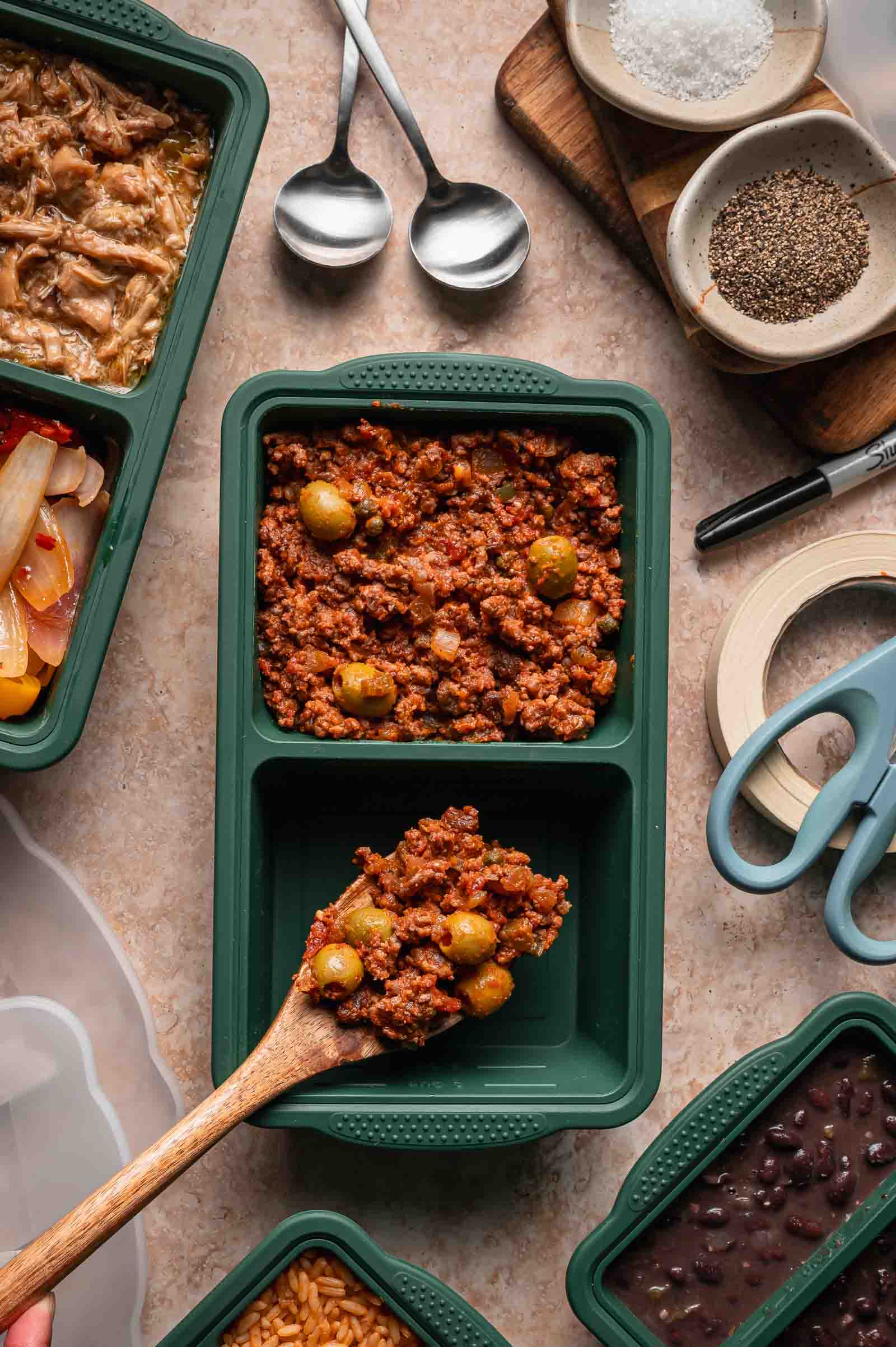 A wooden spoon portions picadillo into a green meal prep container, surrounded by containers of rice, beans, and other ingredients on a countertop.