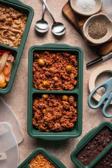 Silicon meal prep container filled with Cuban picadillo, surrounded by cutlery, spices, scissors, rice, and beans on a countertop.