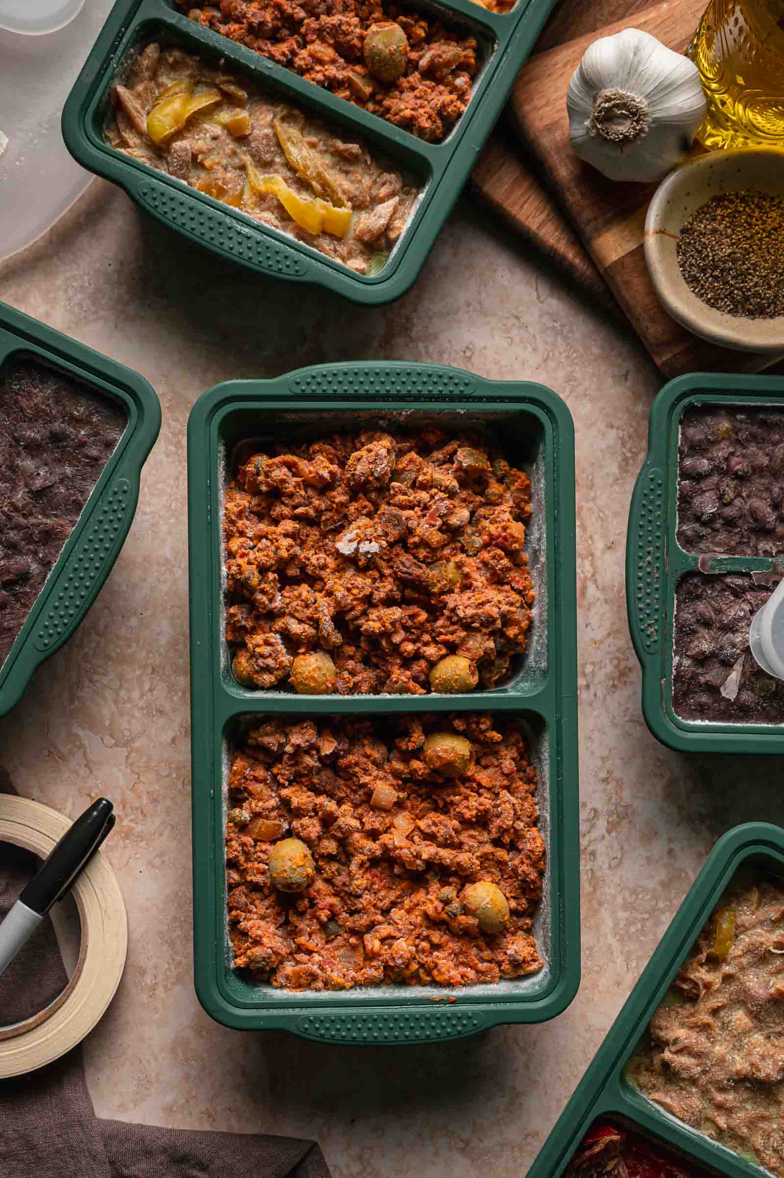 A green meal prep container holds cooked picasdillo, surrounded by similar containers with other prepared meals on a kitchen countertop.