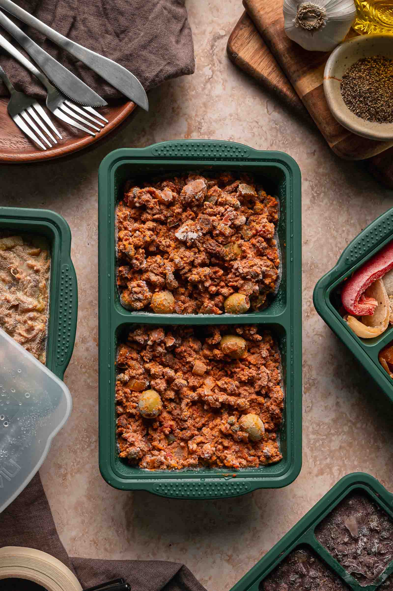 A green meal prep container filled with frozen picadollo sits on a countertop surrounded by utensils and other meal containers.