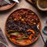 A wooden bowl with black beans, picadillo, and sautéed peppers and onions sits on a table with utensils and various ingredients nearby.