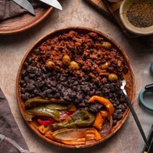 A wooden bowl with black beans, picadillo, and sautéed peppers and onions sits on a table with utensils and various ingredients nearby.