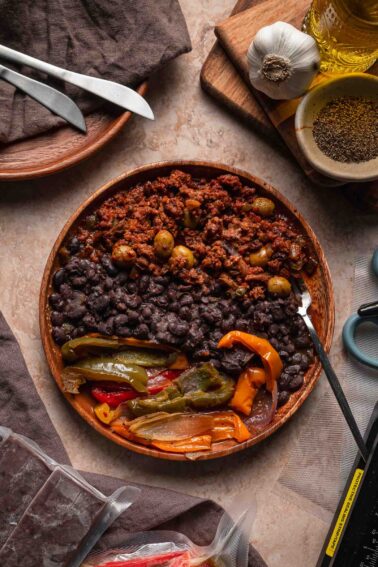 A wooden bowl with black beans, picadillo, and sautéed peppers and onions sits on a table with utensils and various ingredients nearby.