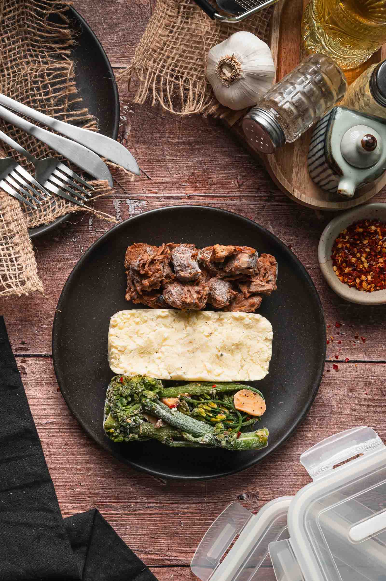 A black plate with frozen adobo pork, mashed potatoes, and broccolini on a wooden table, surrounded by utensils, spices, and kitchen items.