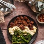A plate with Boursin mashed potatoes, adobo pork, and lemon garlic broccolini on a wooden table with utensils, condiments, and seasonings nearby.