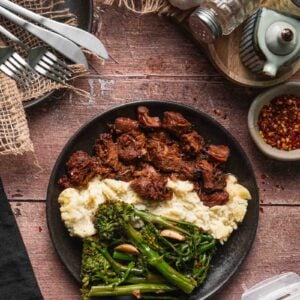 A plate with Boursin mashed potatoes, adobo pork, and lemon garlic broccolini on a wooden table with utensils, condiments, and seasonings nearby.