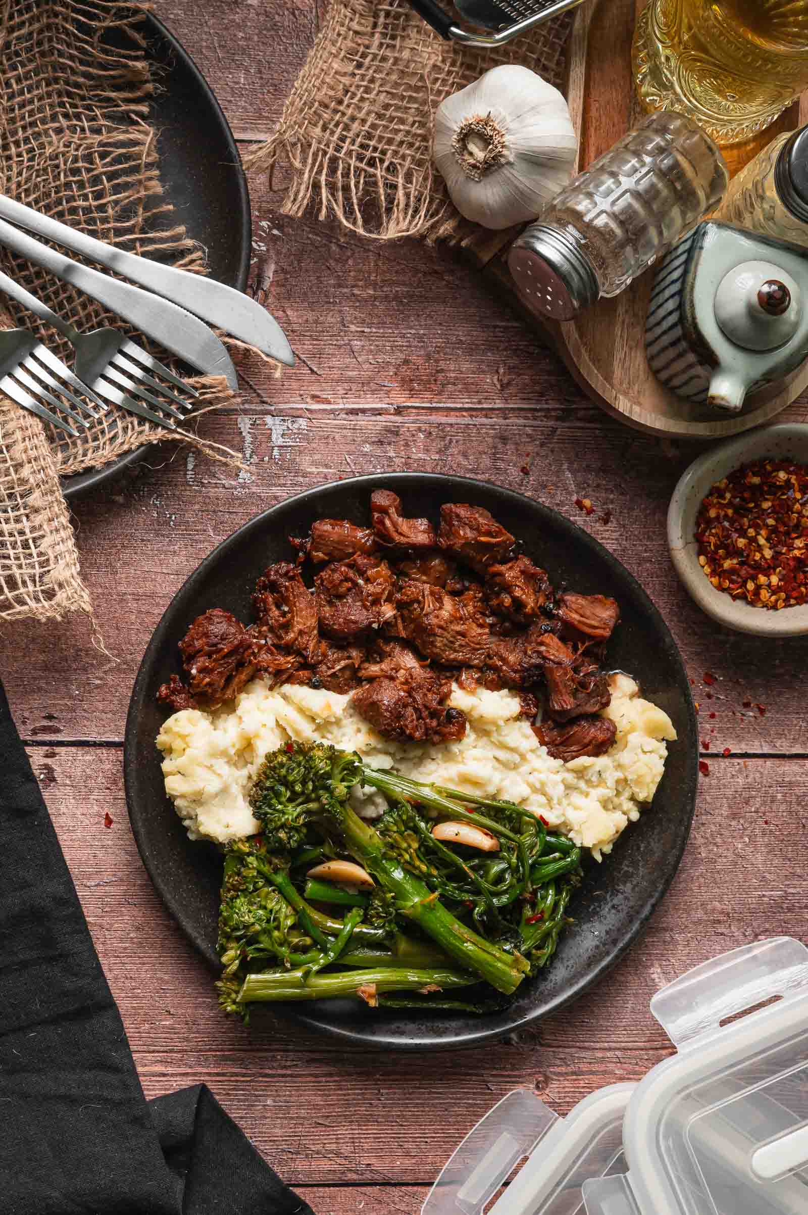 A plate with Boursin mashed potatoes, adobo pork, and lemon garlic broccolini on a wooden table with utensils, condiments, and seasonings nearby.