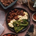 A plate with Boursin mashed potatoes, pork adobo, and and cooked broccolini sits on a wooden table surrounded by storage containers and seasonings.