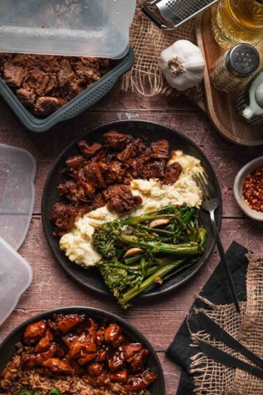 A plate with Boursin mashed potatoes, pork adobo, and and cooked broccolini sits on a wooden table surrounded by storage containers and seasonings.
