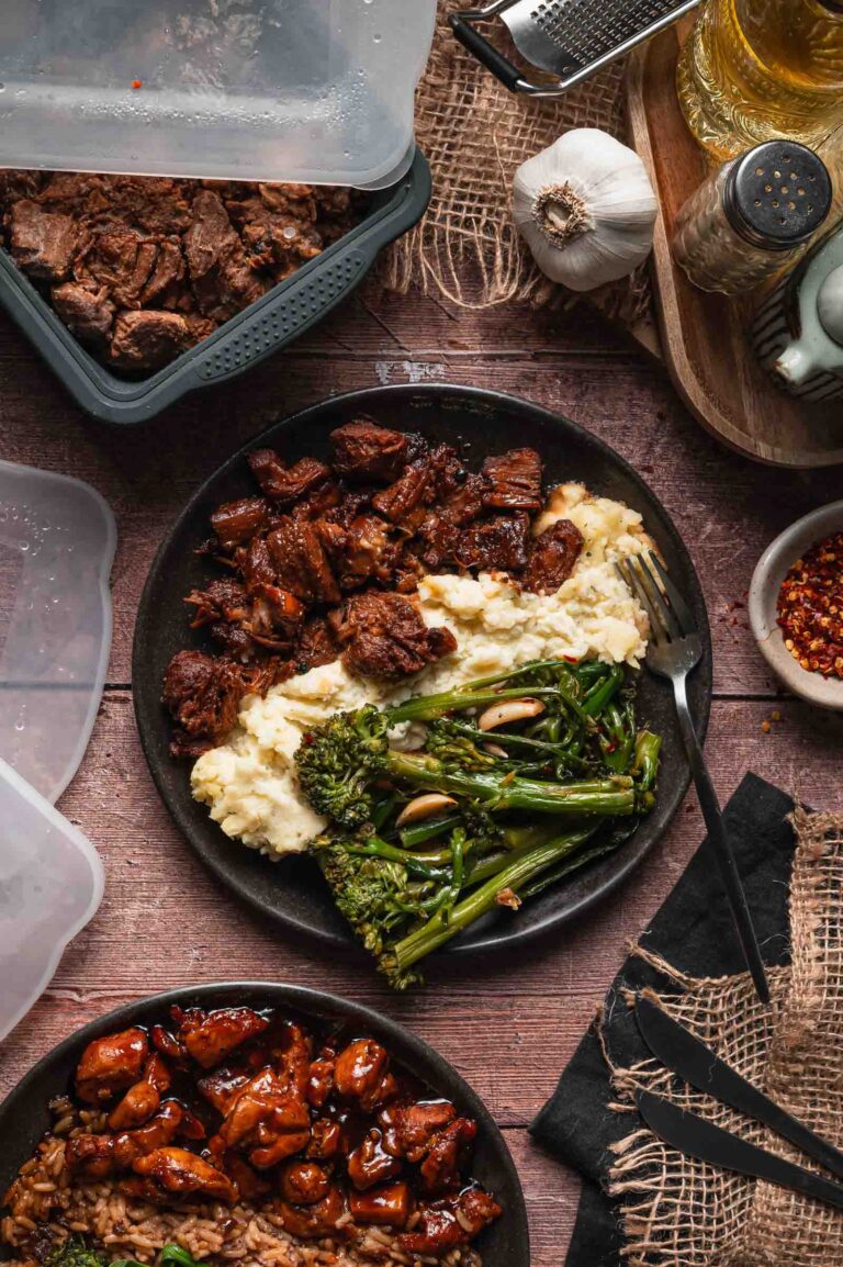 A plate with Boursin mashed potatoes, pork adobo, and and cooked broccolini sits on a wooden table surrounded by storage containers and seasonings.