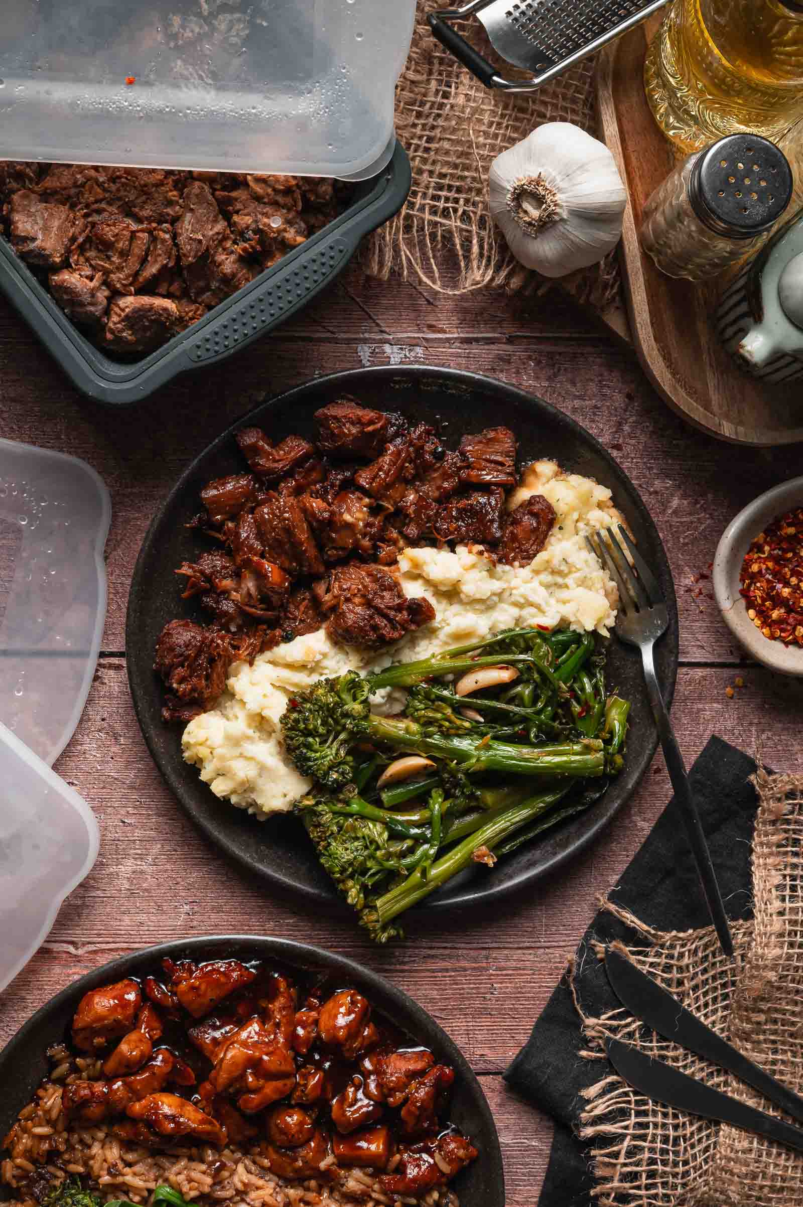 A plate with Boursin mashed potatoes, pork adobo, and and cooked broccolini sits on a wooden table surrounded by storage containers and seasonings.