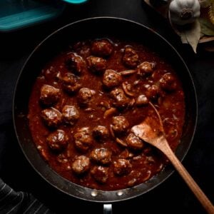 A skillet filled with salisbury steak meatballs in sauce sits on a dark surface with a wooden spoon; garlic and spices are nearby.