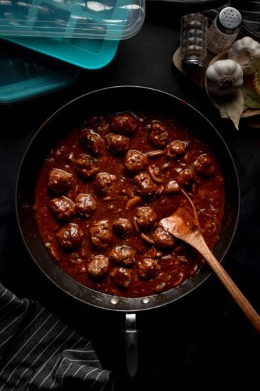 A skillet filled with salisbury steak meatballs in sauce sits on a dark surface with a wooden spoon; garlic and spices are nearby.