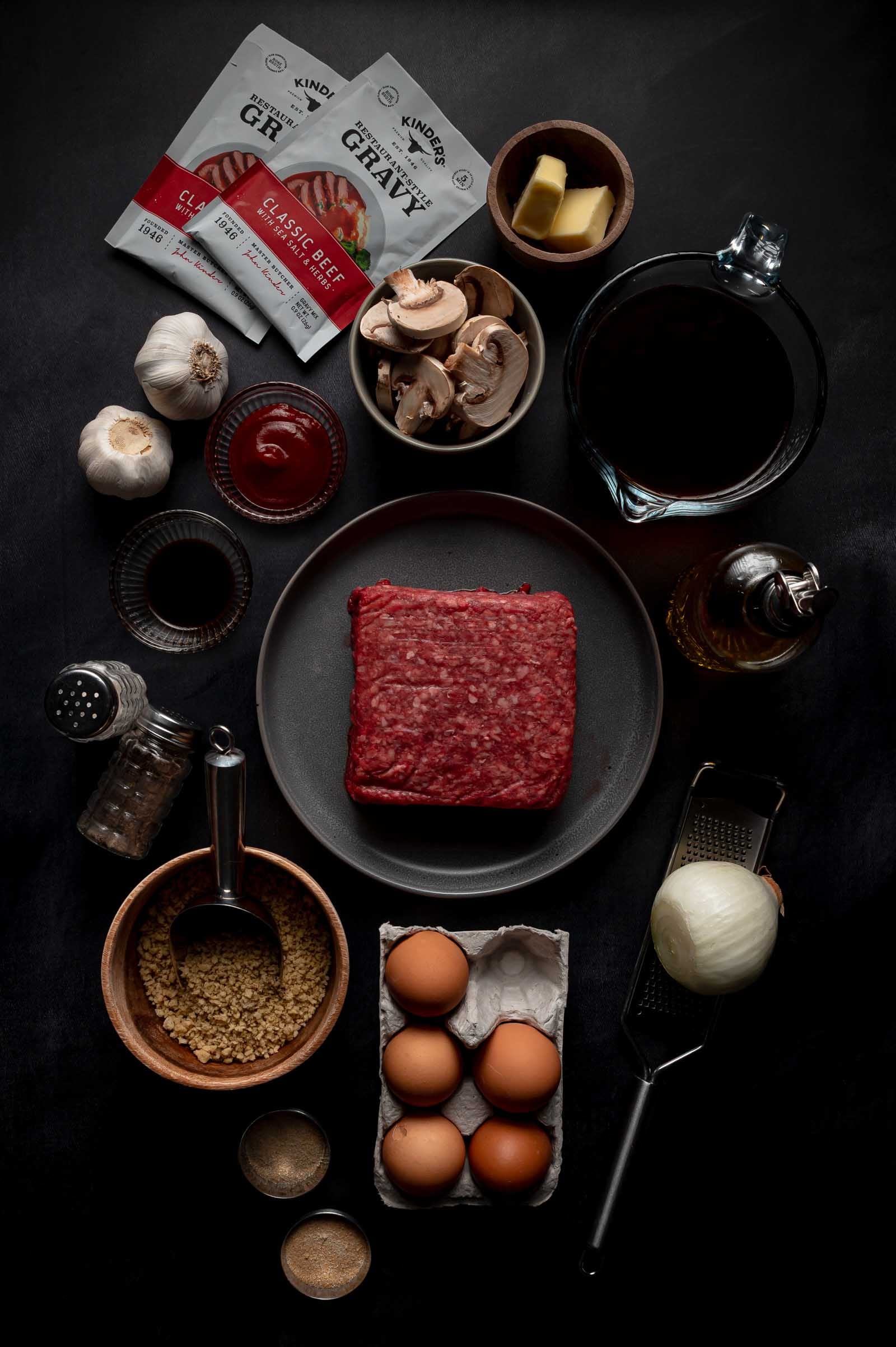 Top-down view of meatball ingredients on a dark surface, including ground beef, eggs, onion, mushrooms, garlic, gravy packets, ketchup, butter, broth, oil, spices, and breadcrumbs.