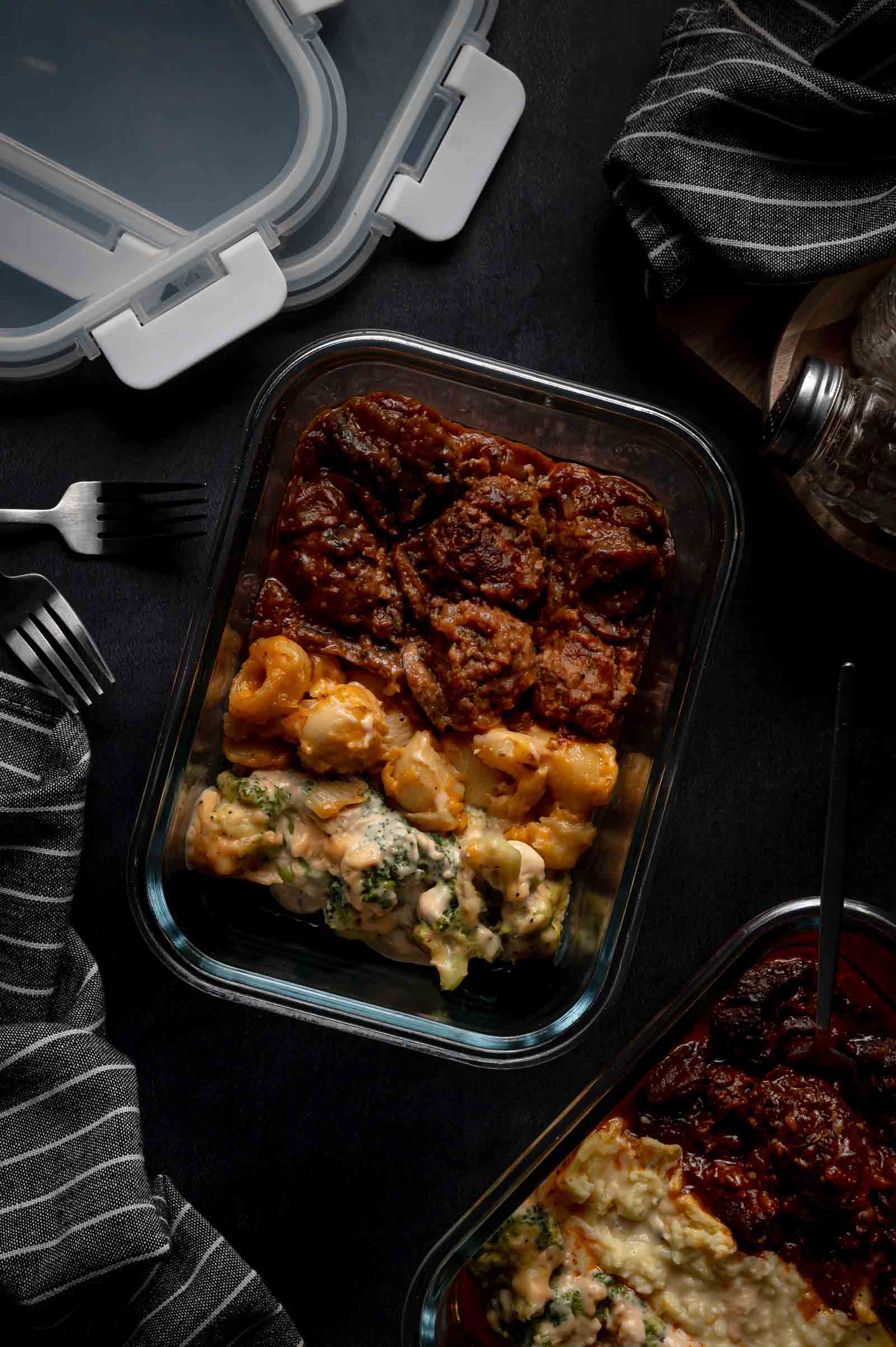 Glass container with mac and cheese, meatballs and broccili on a dark table, surrounded by striped napkins, cutlery, and a plastic container lid.