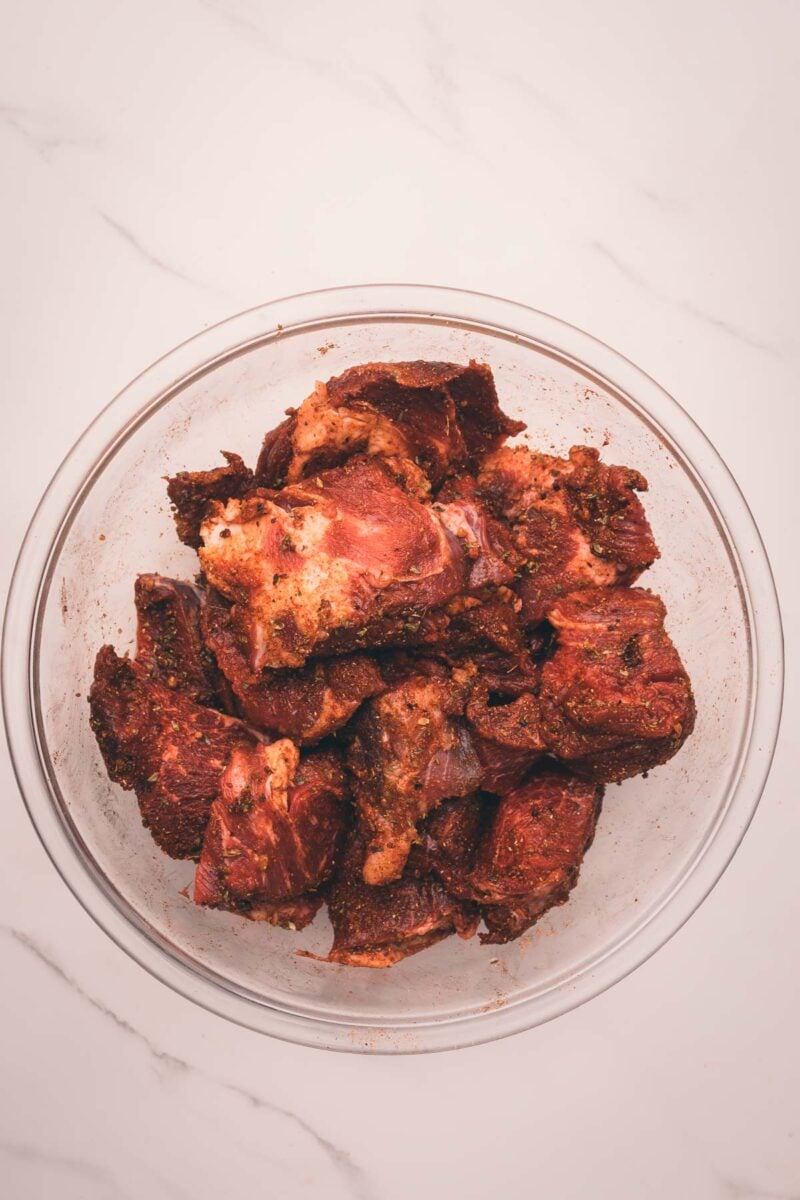 A glass bowl filled with chunks of raw, seasoned meat sits on a white marble surface.