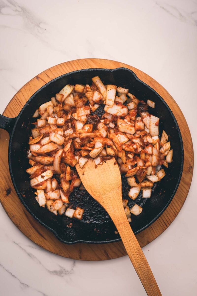 Diced onions with spices are being sautéed in a black cast iron skillet coated in caramelized tomato paste.