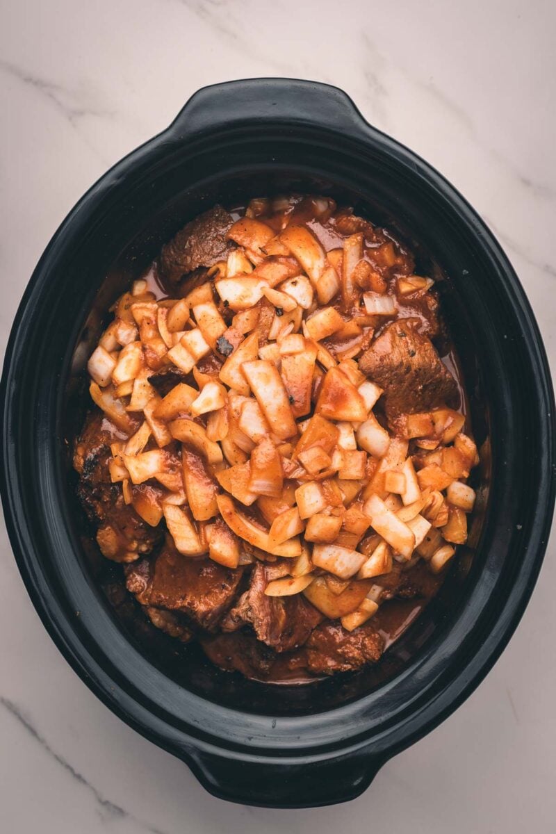 Chunks of raw beef and chopped onions in a black slow cooker, coated with a sauce, ready for cooking.