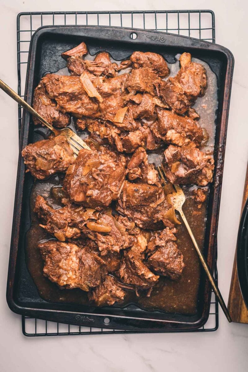 Cooked pieces of slow cooker Mexican beef with sauce on a black baking tray, with two serving forks, placed on a cooling rack over a white surface.