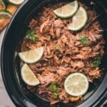 Shredded Mexican beef in a slow cooker, garnished with lime slices and cilantro, with extra lime slices on a cutting board nearby.