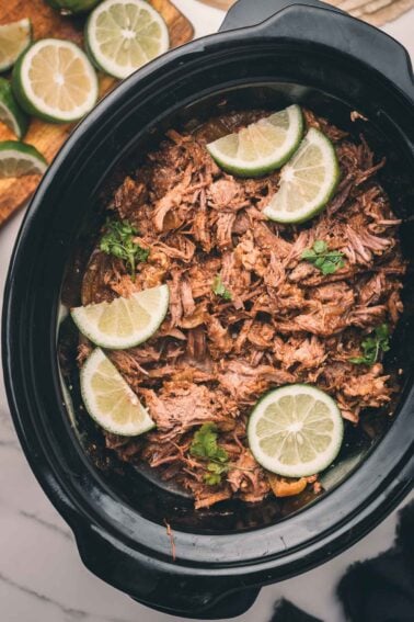 Shredded Mexican beef in a slow cooker, garnished with lime slices and cilantro, with extra lime slices on a cutting board nearby.