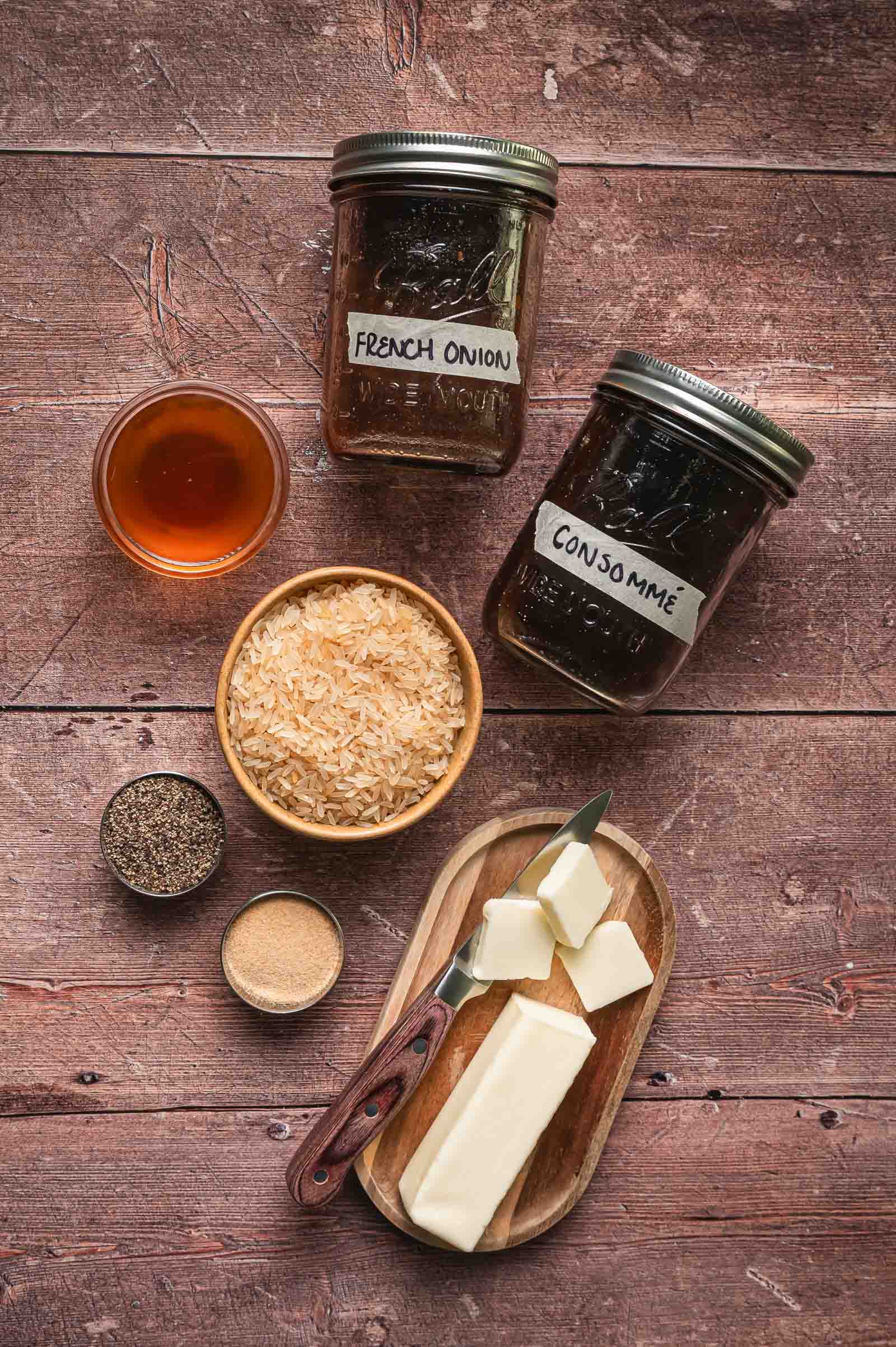 Overhead view of ingredients for stick of butter rice on a wooden surface: jars labeled "French Onion" and "Consommé," a bowl of rice, butter, a knife, black pepper, and seasoning.