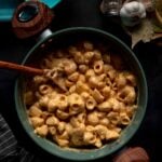 A pot of creamy macaroni and cheese with a wooden spoon, surrounded by garlic, spices, bay leaves, and storage containers on a dark surface.