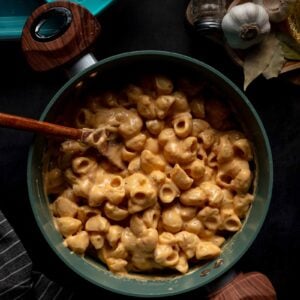 A pot of creamy macaroni and cheese with a wooden spoon, surrounded by garlic, spices, bay leaves, and storage containers on a dark surface.