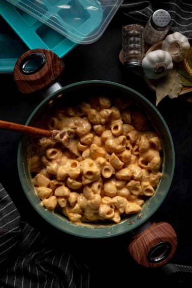 A pot of creamy macaroni and cheese with a wooden spoon, surrounded by garlic, spices, bay leaves, and storage containers on a dark surface.