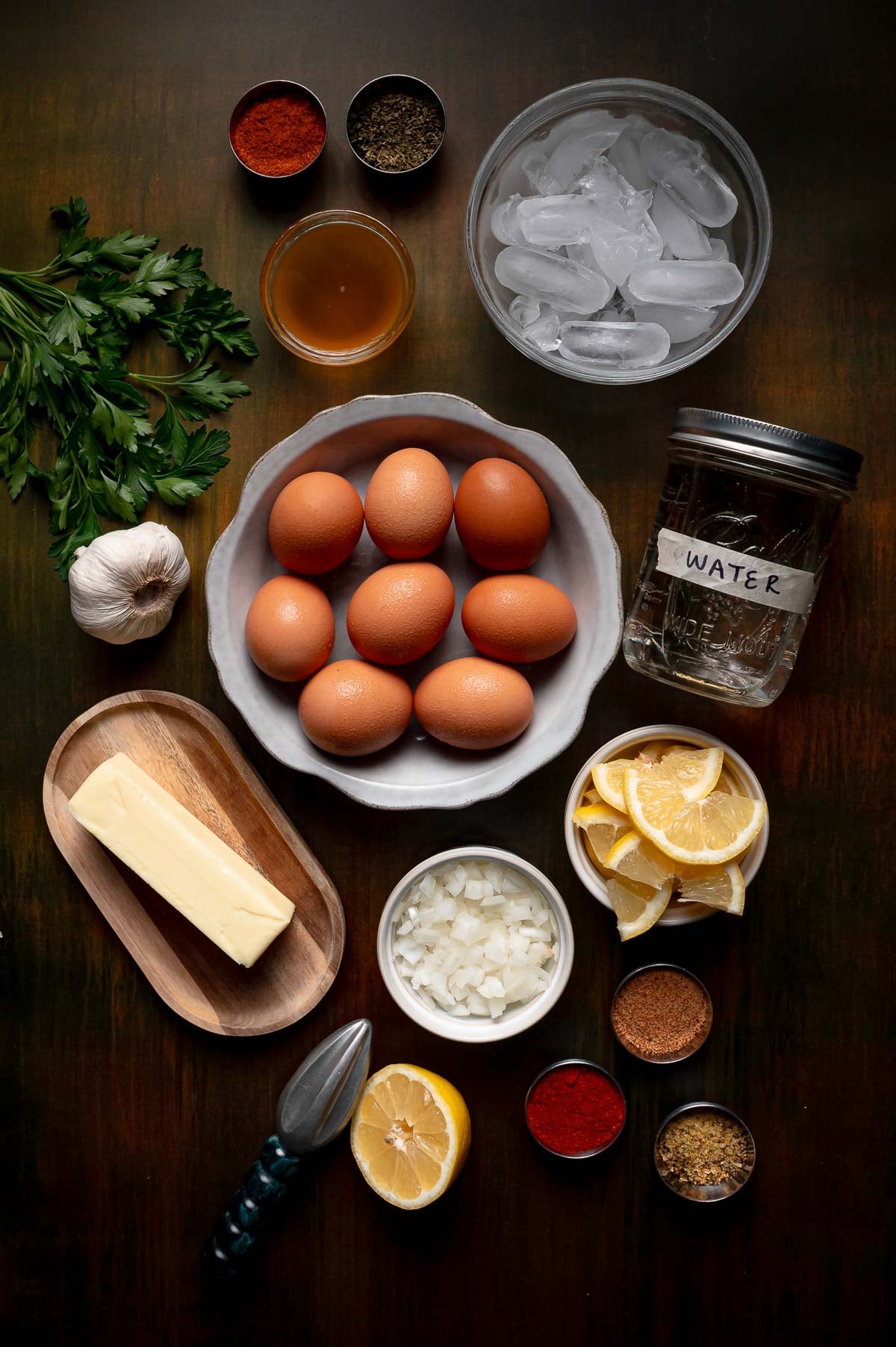 A flat lay of eggs, butter, chopped onion, garlic, parsley, spices, sliced lemon, ice water, and a citrus juicer arranged on a dark wooden surface.