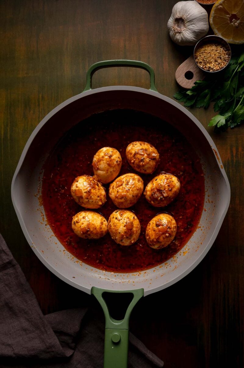 Eight hard-boiled eggs coated in a red spiced sauce in a skillet. Garlic, parsley, a lemon half, and spices are placed nearby on the wooden surface.