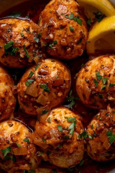 Boiled eggs coated in a spiced red sauce, garnished with chopped herbs, and served with lemon wedges.