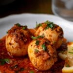 Three hard-boiled eggs covered in spicy red sauce, garnished with chopped herbs, served on a white plate with a lemon wedge.