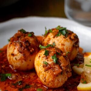 Three hard-boiled eggs covered in spicy red sauce, garnished with chopped herbs, served on a white plate with a lemon wedge.