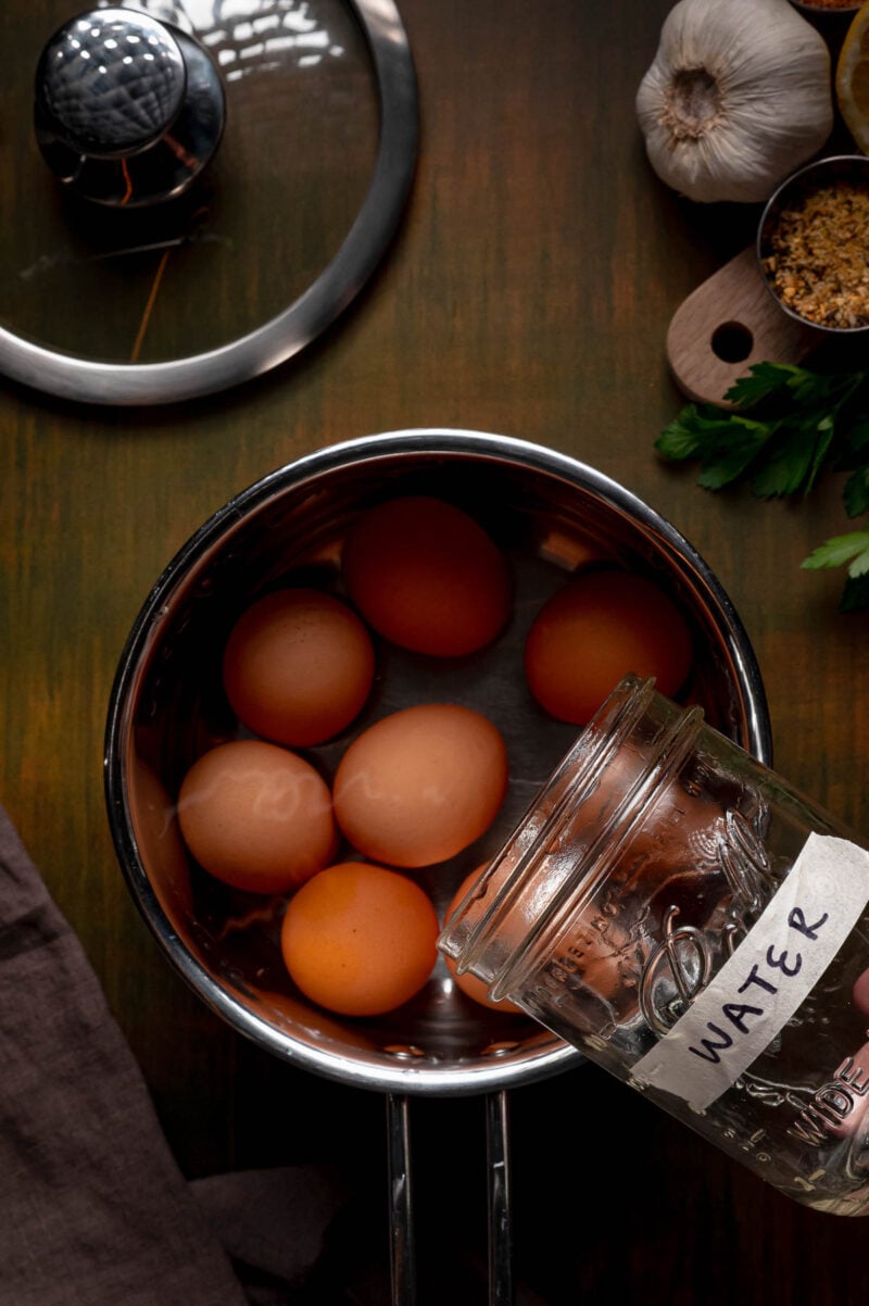 A pot with brown eggs inside sits on a wooden surface while a hand pours water from a labeled jar. Garlic, parsley, and brown sugar are nearby.