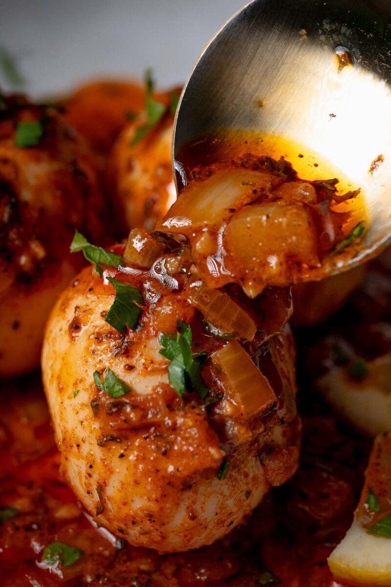 A close-up of hard-boiled eggs coated in a rich, spiced sauce with onions and herbs, being spooned over with extra sauce.