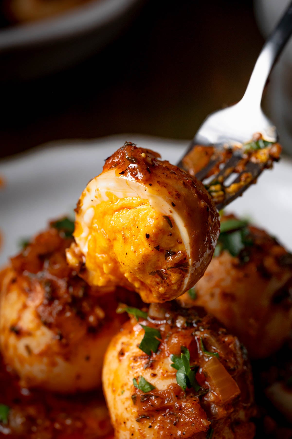 A fork holds a halved boiled egg coated in spicy red sauce, with other sauced eggs and chopped herbs on a plate below.