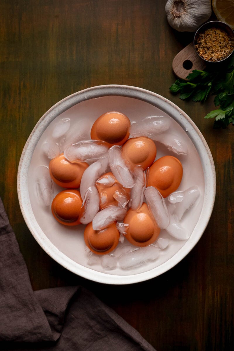 A bowl of brown eggs sits in an ice water bath with ice cubes. Parsley, brown sugar, and garlic are nearby on a wooden surface.