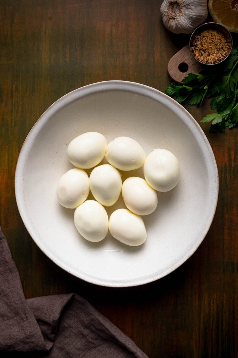 A white bowl containing peeled hard-boiled eggs, placed on a wooden surface with garlic, parsley, brown sugar, and a lemon wedge nearby.