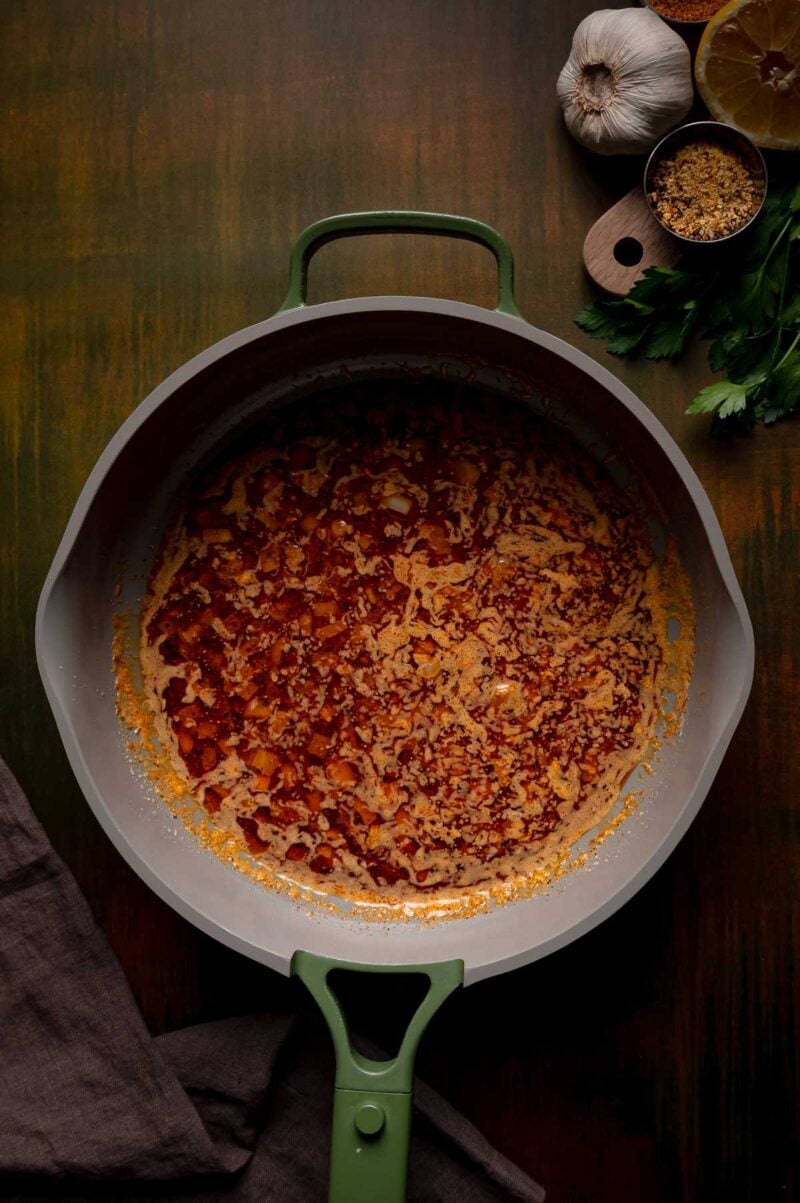 A large skillet with a spicy butter mixture, surrounded by garlic, parsley, lemon, and a measuring cup with spices.