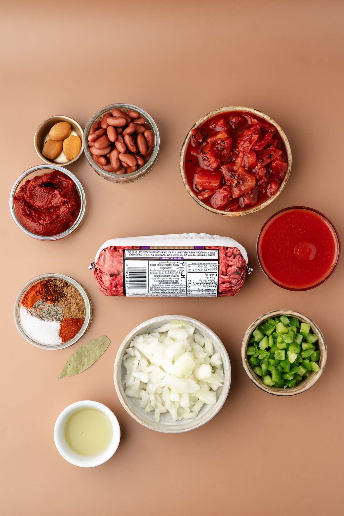 Various chili ingredients on a tan surface, including ground beef, chopped onion, green pepper, kidney beans, tomatoes, tomato sauce, tomato paste, spices, garlic, and a small bowl of oil.