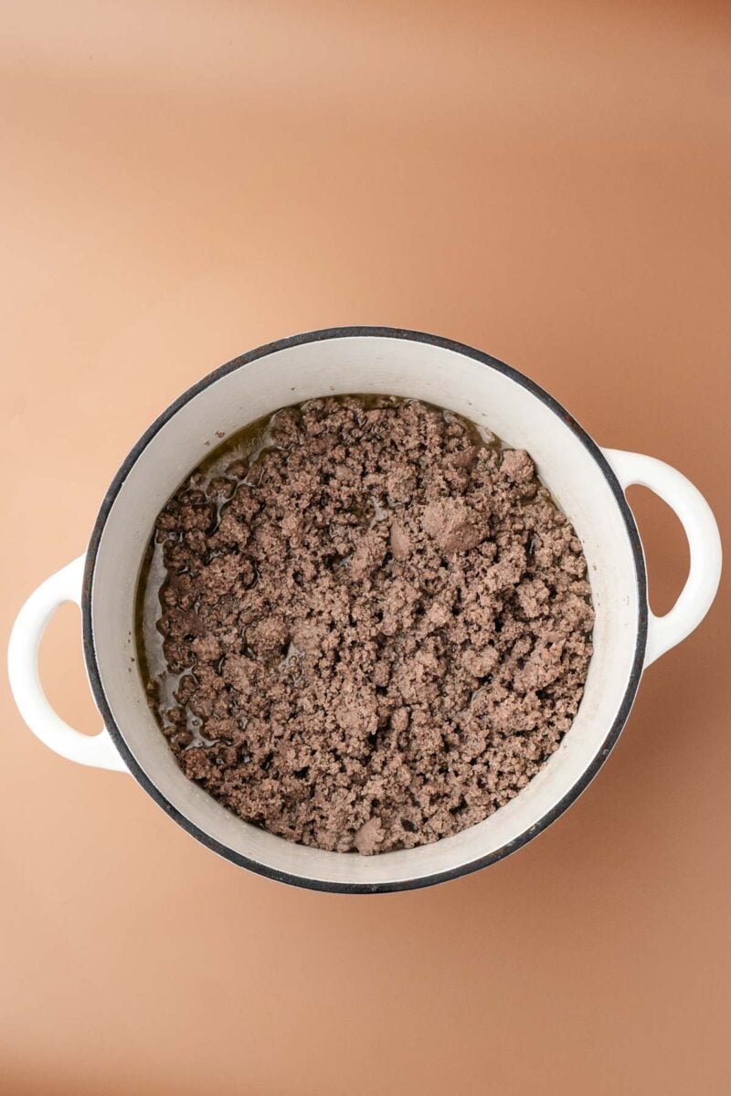 Cooked ground beef in a white Dutch oven with handles, viewed from above, on a tan surface.