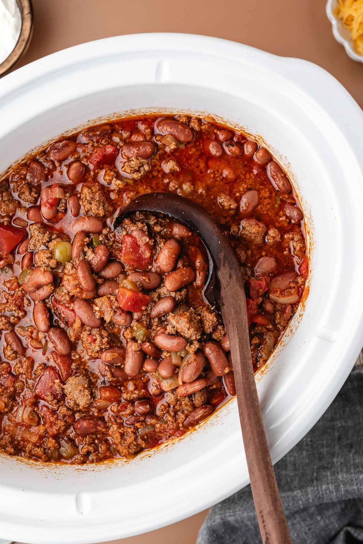 A slow cooker filled with chili made of ground meat, red beans, tomatoes, and spices, with a wooden ladle resting inside.