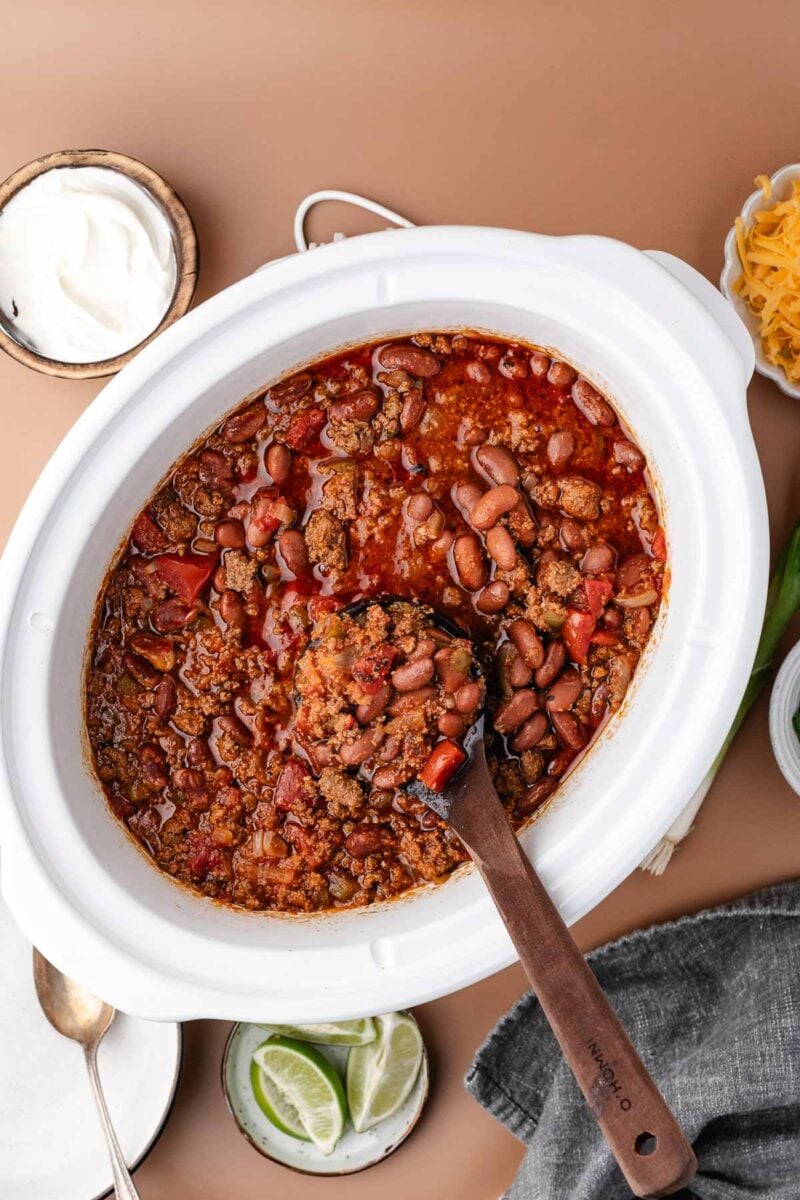 A white slow cooker filled with chili made of ground beef, beans, and tomatoes. A wooden spoon rests inside. Surrounding items include sour cream, shredded cheese, and lime slices.
