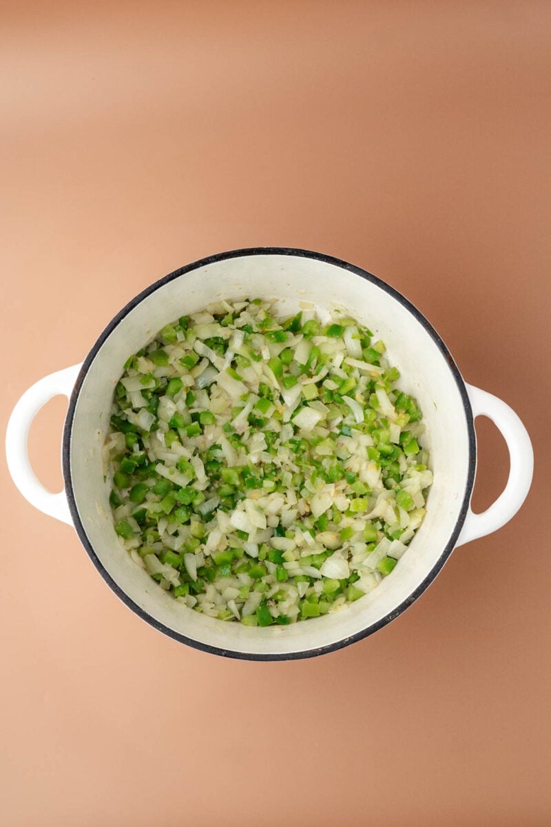 White pot filled with sautéed chopped onions and green bell peppers on a neutral tan background, viewed from above.