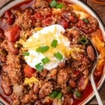 Bowl of chili with ground meat, beans, and tomatoes, topped with shredded cheese, sour cream, and chopped green onions, with a spoon resting in the bowl.