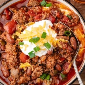 Bowl of chili with ground meat, beans, and tomatoes, topped with shredded cheese, sour cream, and chopped green onions, with a spoon resting in the bowl.
